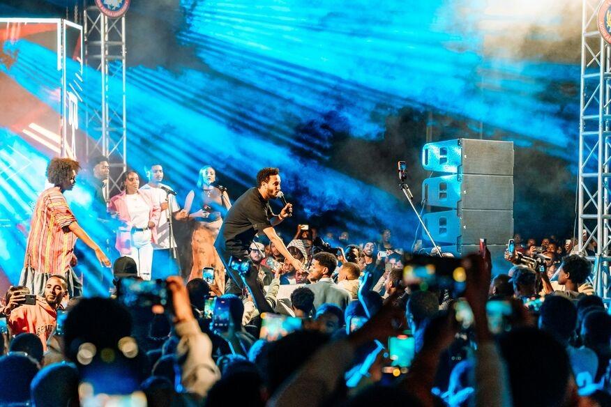 A vibrant concert scene with a singer engaging the lively crowd under blue stage lights. Audience members record the moment with their phones, capturing the energetic atmosphere.