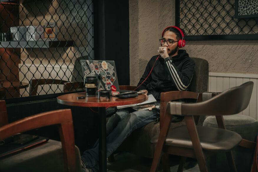 Person wearing red headphones, deep in thought, sits in a cozy café. A laptop with stickers rests on the table, creating a studious ambiance.