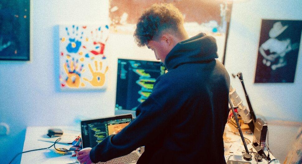 A person in a hoodie works at a desk with two monitors displaying code. The room is warmly lit, featuring colorful handprint art and a black-and-white photo.