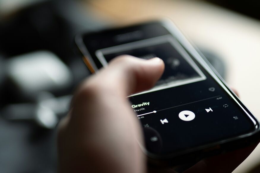 Close-up of a hand holding a smartphone playing music. The screen shows a song titled "Gravity" with playback controls. The mood is relaxed.