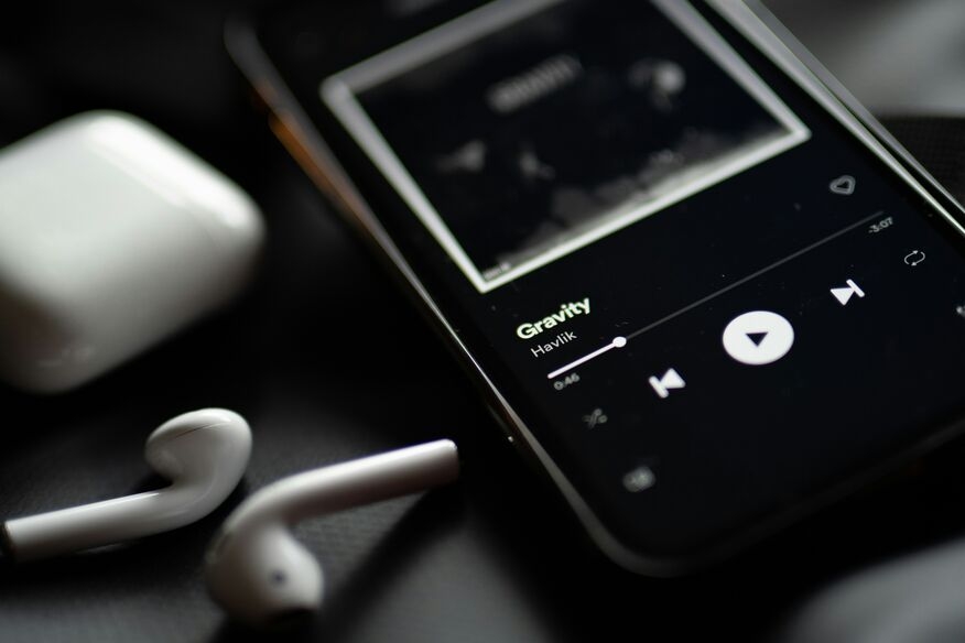 Close-up of a smartphone playing music with the song "Gravity" on screen. White wireless earbuds and a charging case lie nearby on a dark surface.