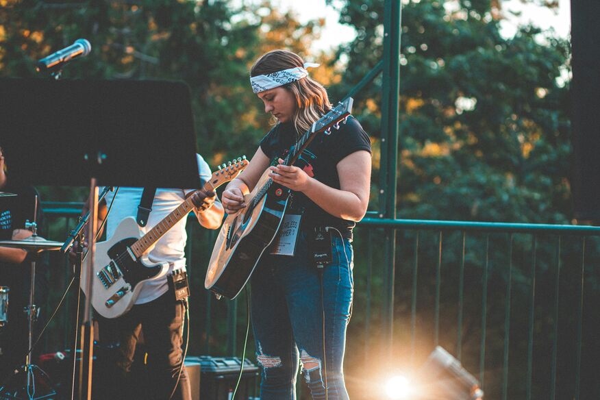 Young woman playing acoustic guitar on an outdoor stage, wearing a bandana and jeans, surrounded by bandmates, with trees in the background and warm lighting.
