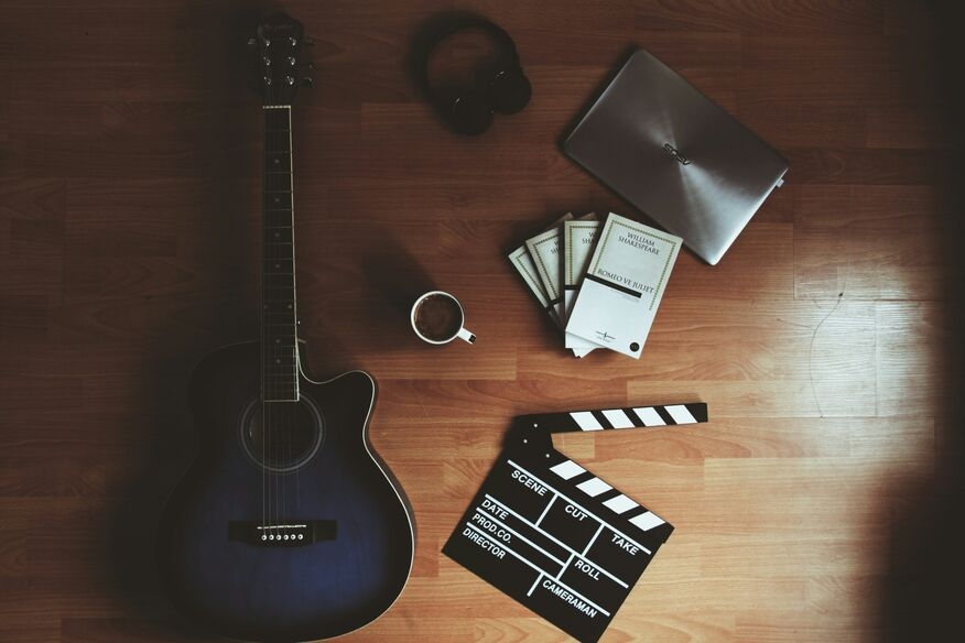 Aerial view of a wooden floor with a blue acoustic guitar, film clapperboard, stack of books, laptop, headphones, and a coffee cup, suggesting creativity.